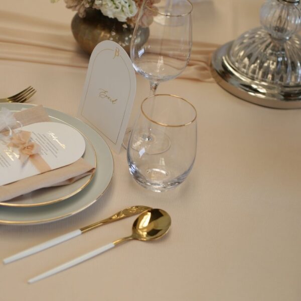 Wit-gouden bestek huren – Feline Styling Wit-gouden bestek naast gestapelde borden en een stoffen servet op een beige tafel