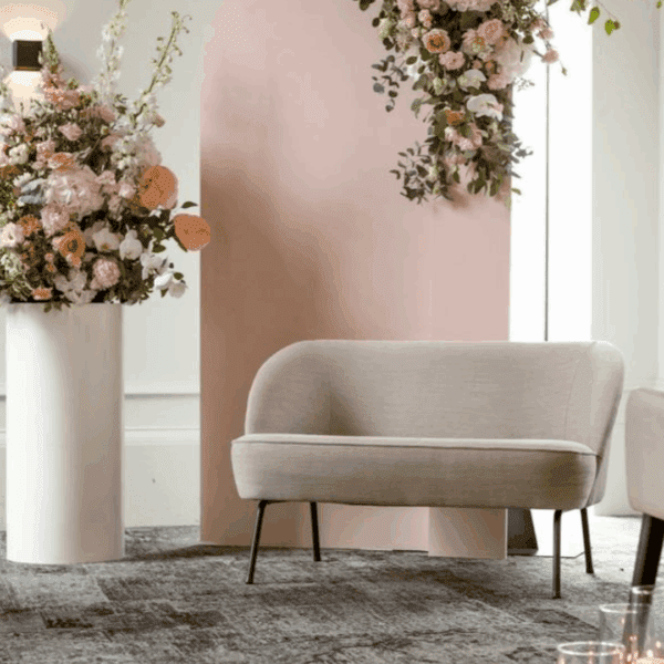Beige trouwbankje – stijlvol ceremoniebankje met ronde vormen Beige trouwbankje met ronde vormen, slanke zwarte poten en zachte bekleding, gestyled met pastelbloemen en een roze backdrop — ideaal voor ceremonie of fotoshoot.