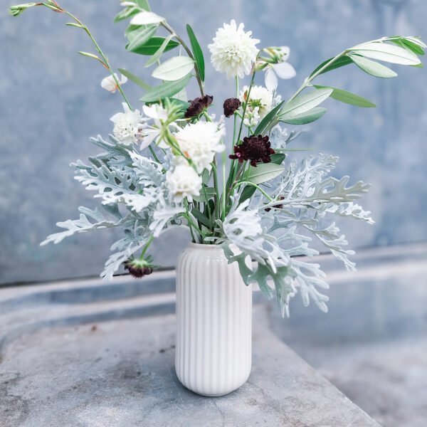 Wit ribbelvaasje met groen en witte bloemen – bruiloft decoratie Wit ribbelvaasje met bloemstuk van witte dahlia’s en groen blad tegen een grijze achtergrond.