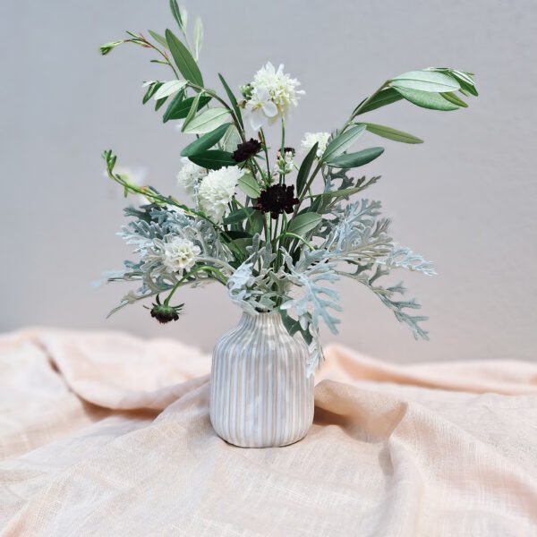 Beige ribbelvaasje met bloemen – verhuur Beige ribbelvaasje met smalle opening, gevuld met groene takken en witte bloemen, te huur bij Feline Styling.
