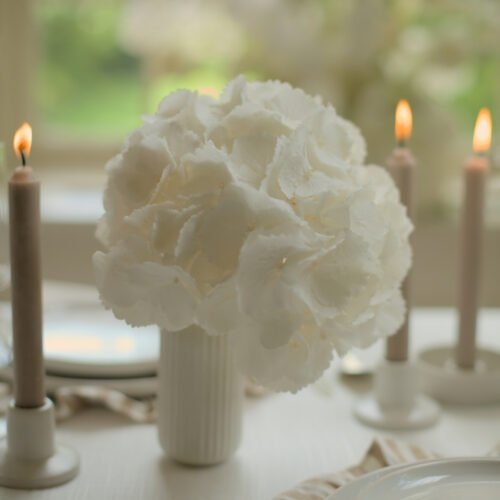 Wit ribbelvaasje met witte hortensia tussen brandende kaarsen, als bruiloft tafelinspiratie