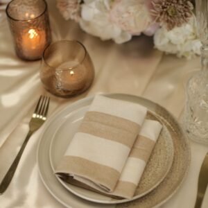 Wit-beige gestreepte servet bij kaarslicht Wit-beige gestreepte servet op een bord, met gouden bestek, amberkleurige windlichten en bloemen op tafel.