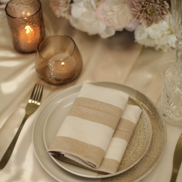 Wit-beige gestreepte servet bij kaarslicht Wit-beige gestreepte servet op een bord, met gouden bestek, amberkleurige windlichten en bloemen op tafel.