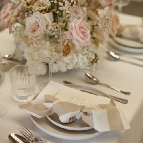 Gedekte tafel met wit-beige gestreepte servetten, glaswerk en een bloemstuk in wit en zachtroze.