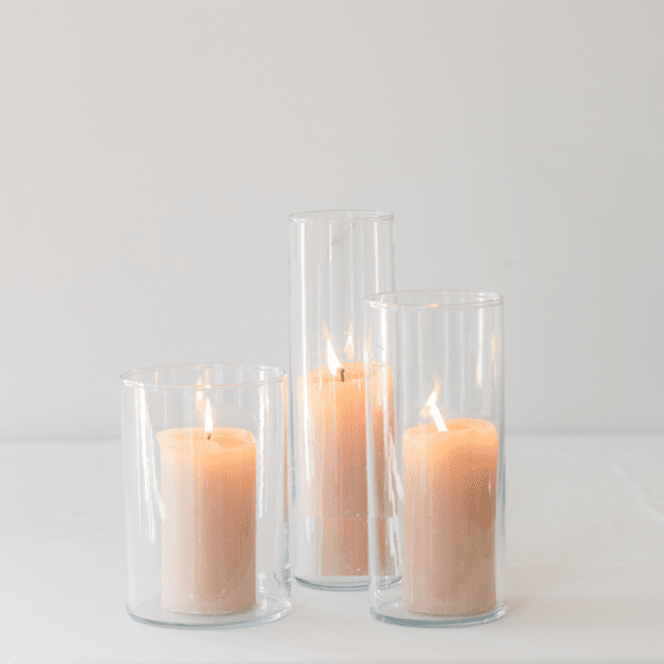 Drie glazen cilinder windlichten met zachte kaarsen in warme neutrale tinten op een rustige lichte ondergrond voor verhuur kaarsenstandaard bruiloft
