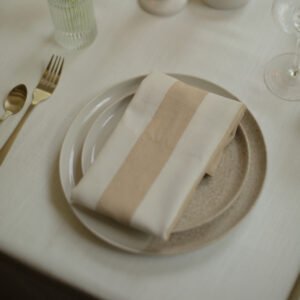 Wit-beige gestreepte servet met gouden bestek Wit-beige gestreepte servet, schuin gevouwen op een licht bord naast gouden bestek.